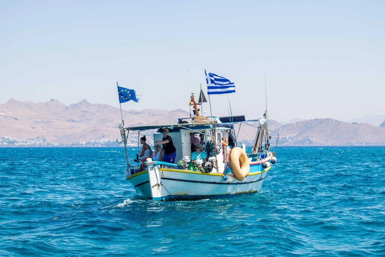De vissersboot met gasten aan dek onder Griekse en Europese vlag, met de bergen van de kust erachter